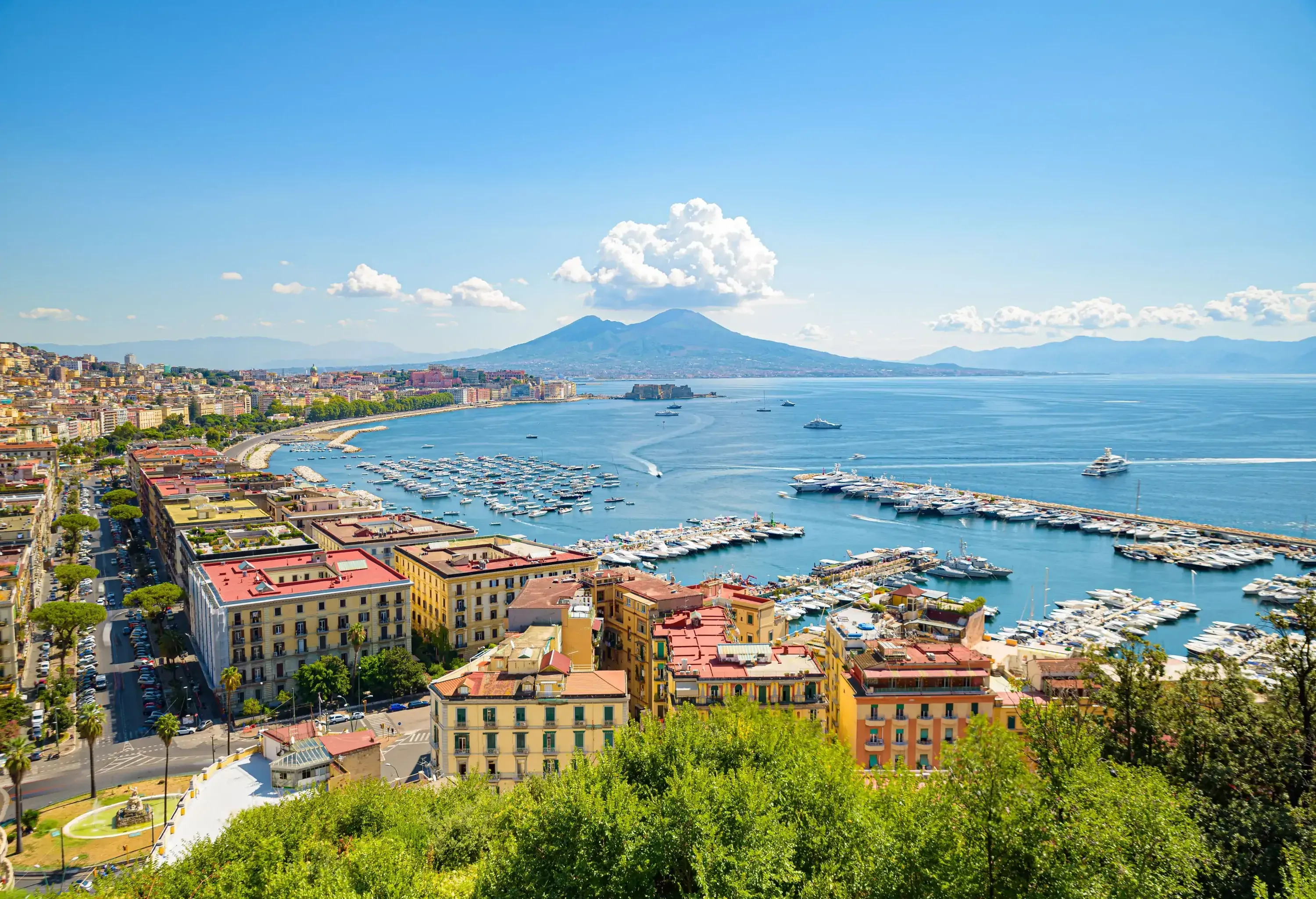 A breathtaking view of a busy harbour in a coastal town, with Mount Vesuvius visible in the distance, overlooking the sea.