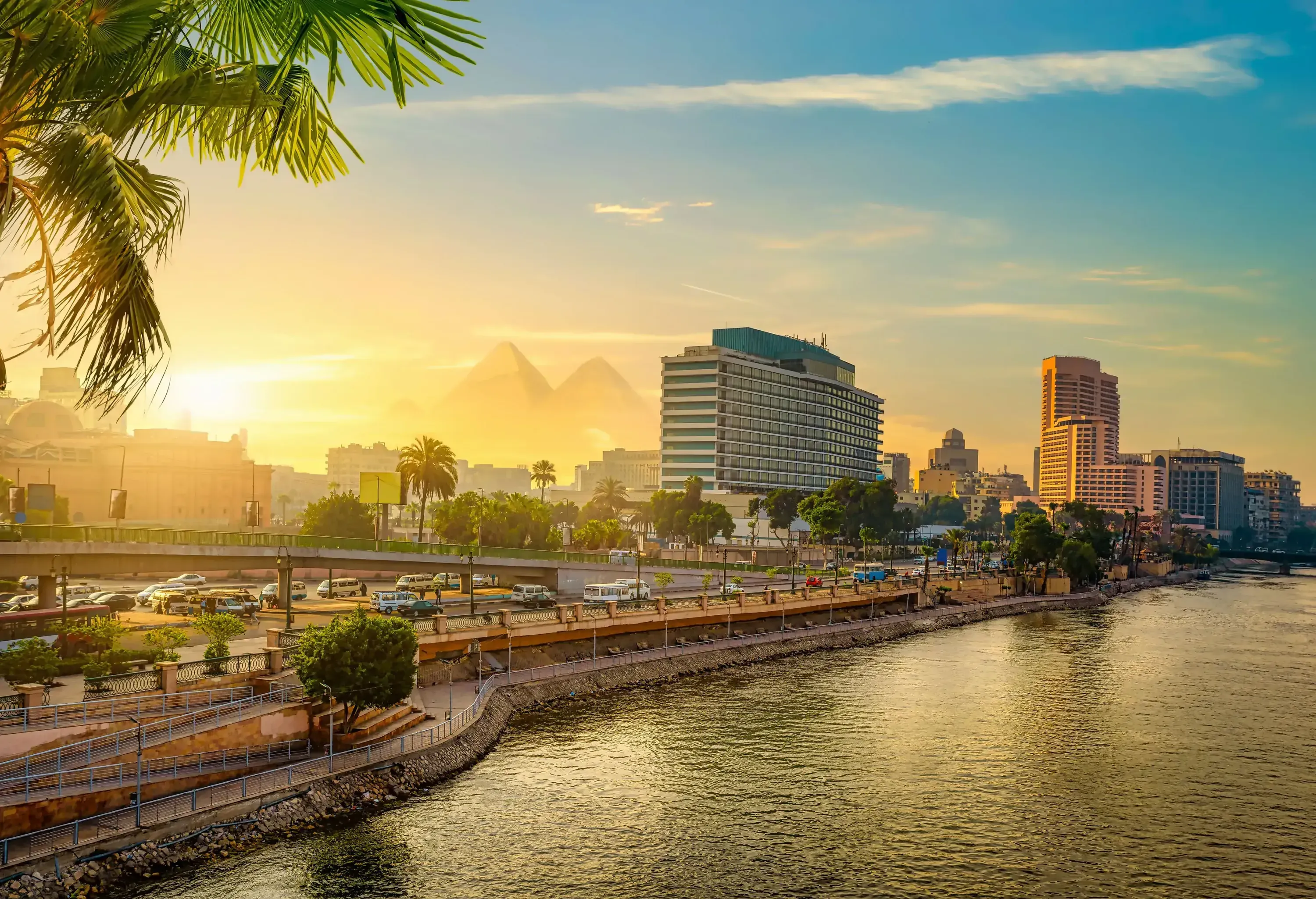 A busy roadway by the river with towering buildings and pyramids in the background.