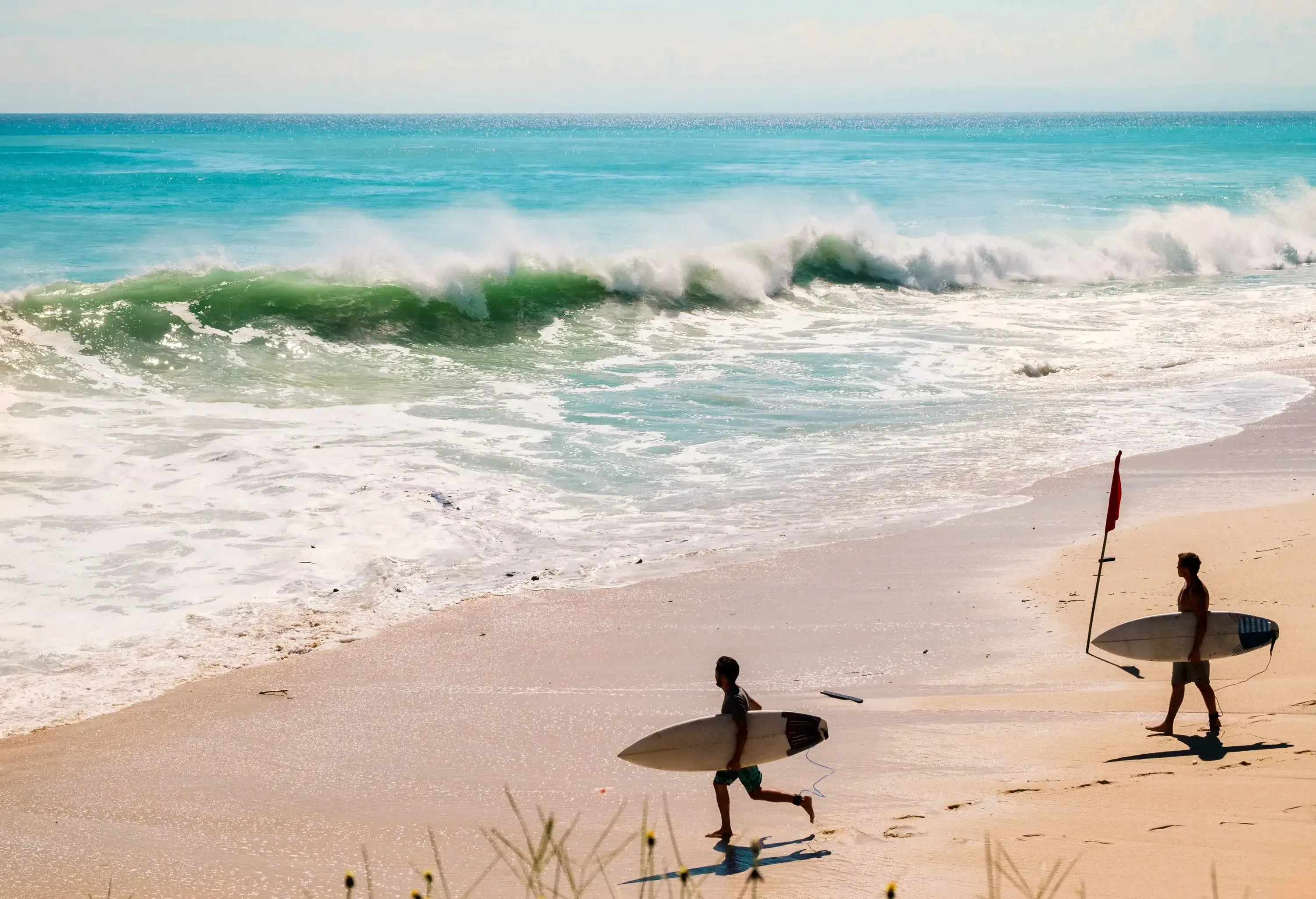 Silhouettes of two surfers carrying their surfboards going towards the massive waves on the beach.