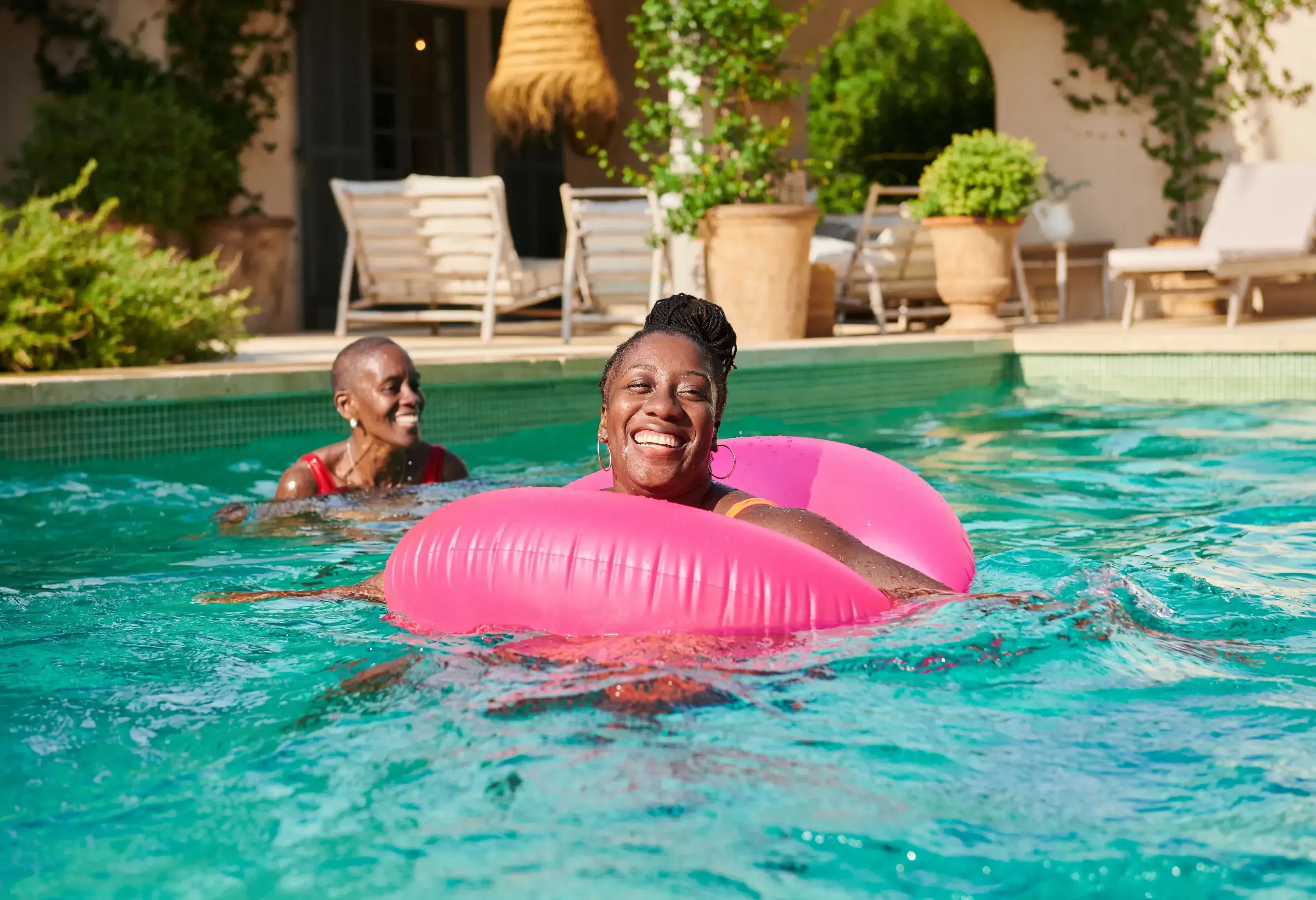 Two people smiling and relaxing in a sunny pool, one on an inflatable ring.