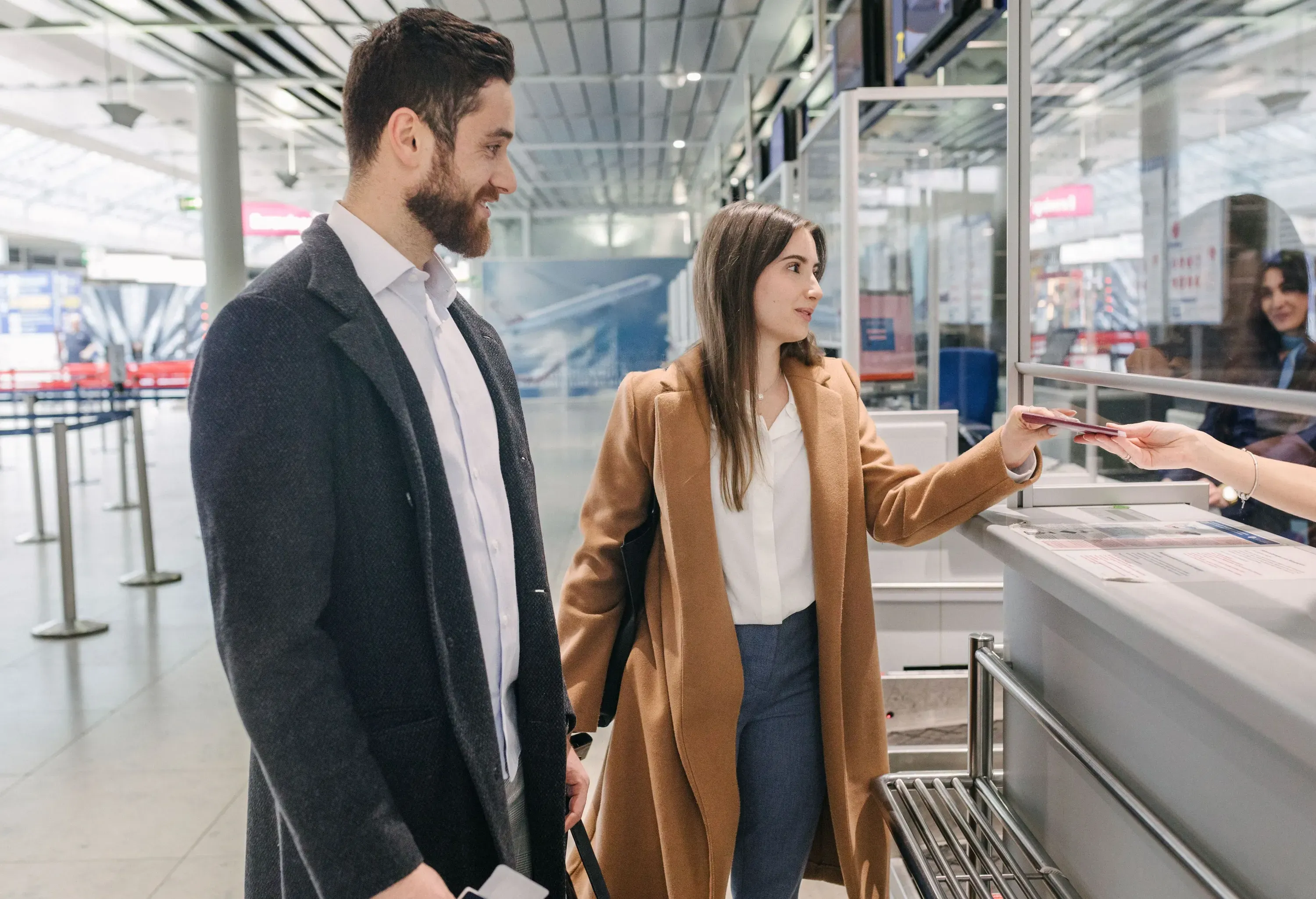 Couple standing in front of an airport terminal, with the woman handing her passport to the person behind the counter.