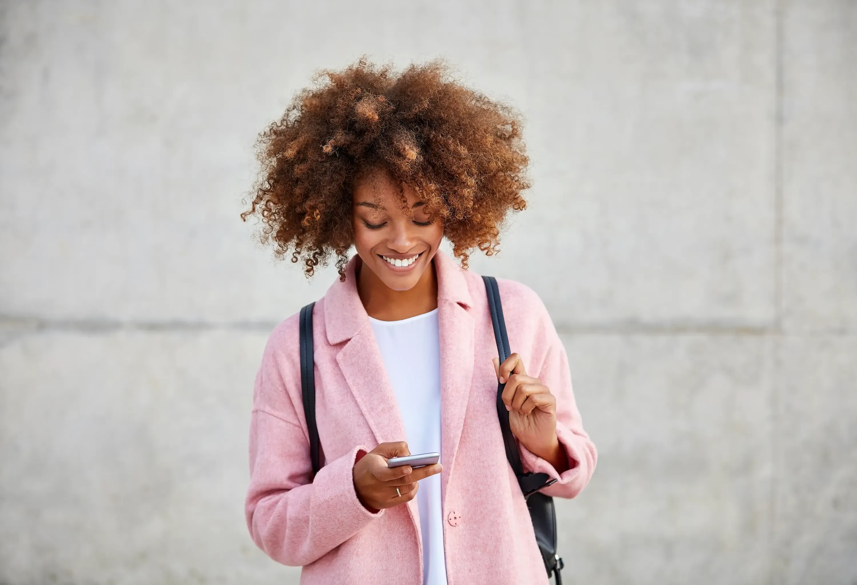 Happy young woman text messaging through smart phone. Beautiful female is with frizzy short hair. She is wearing pink overcoat.