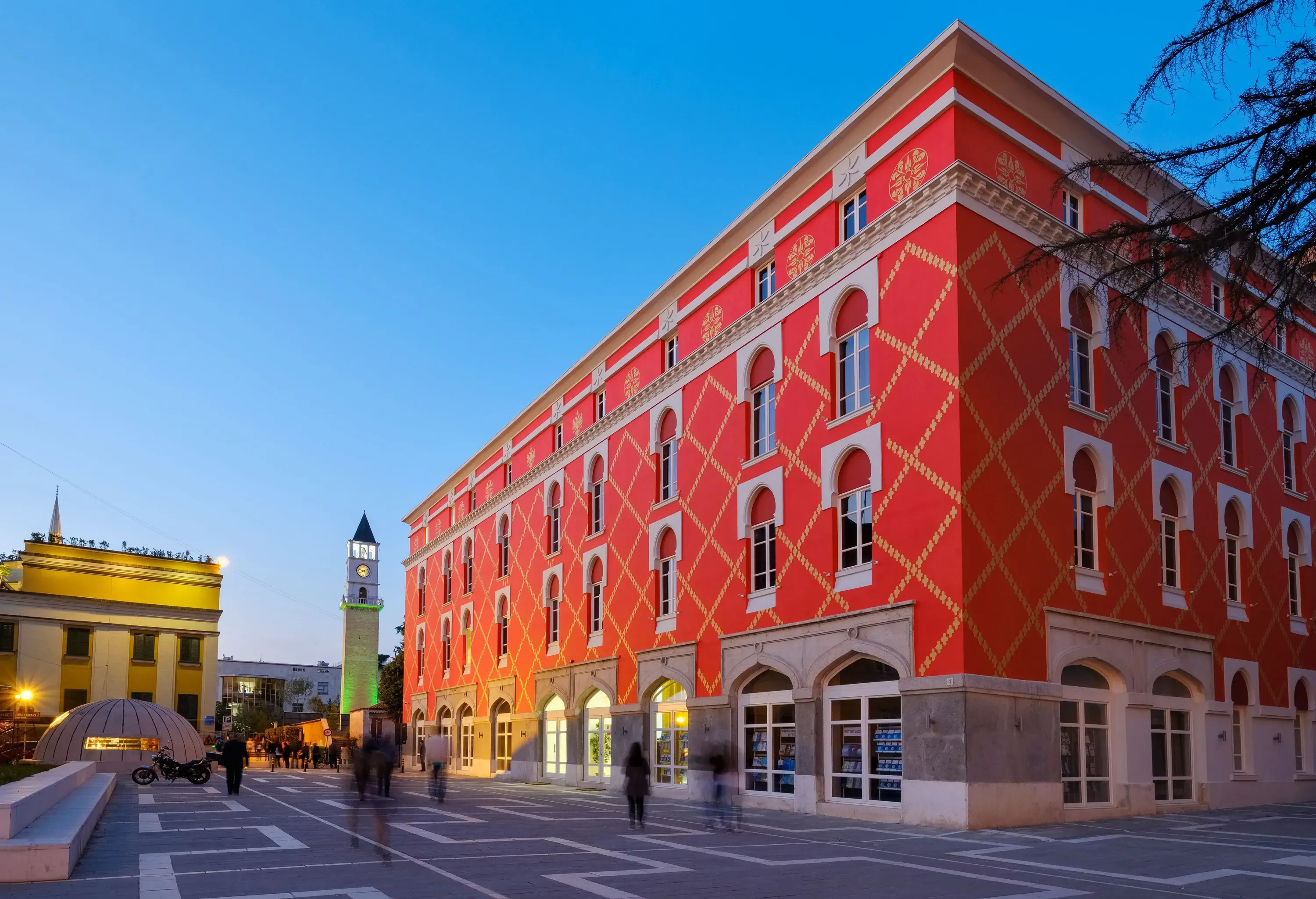 A striking red building with a gold patterned facade stands prominently under a fading sky, with a tall, slender clock tower and other buildings in the distance.
