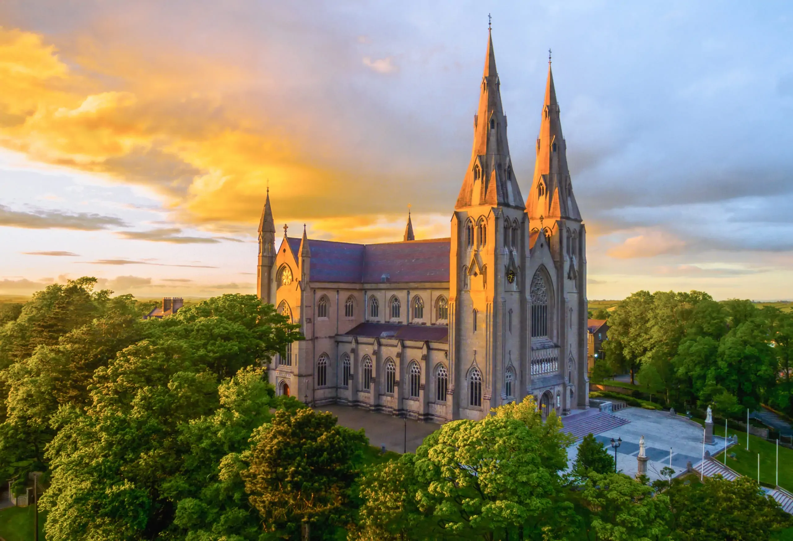 A majestic Gothic revival cathedral with twin spires bathed in the golden light of sunset, surrounded by lush green trees.