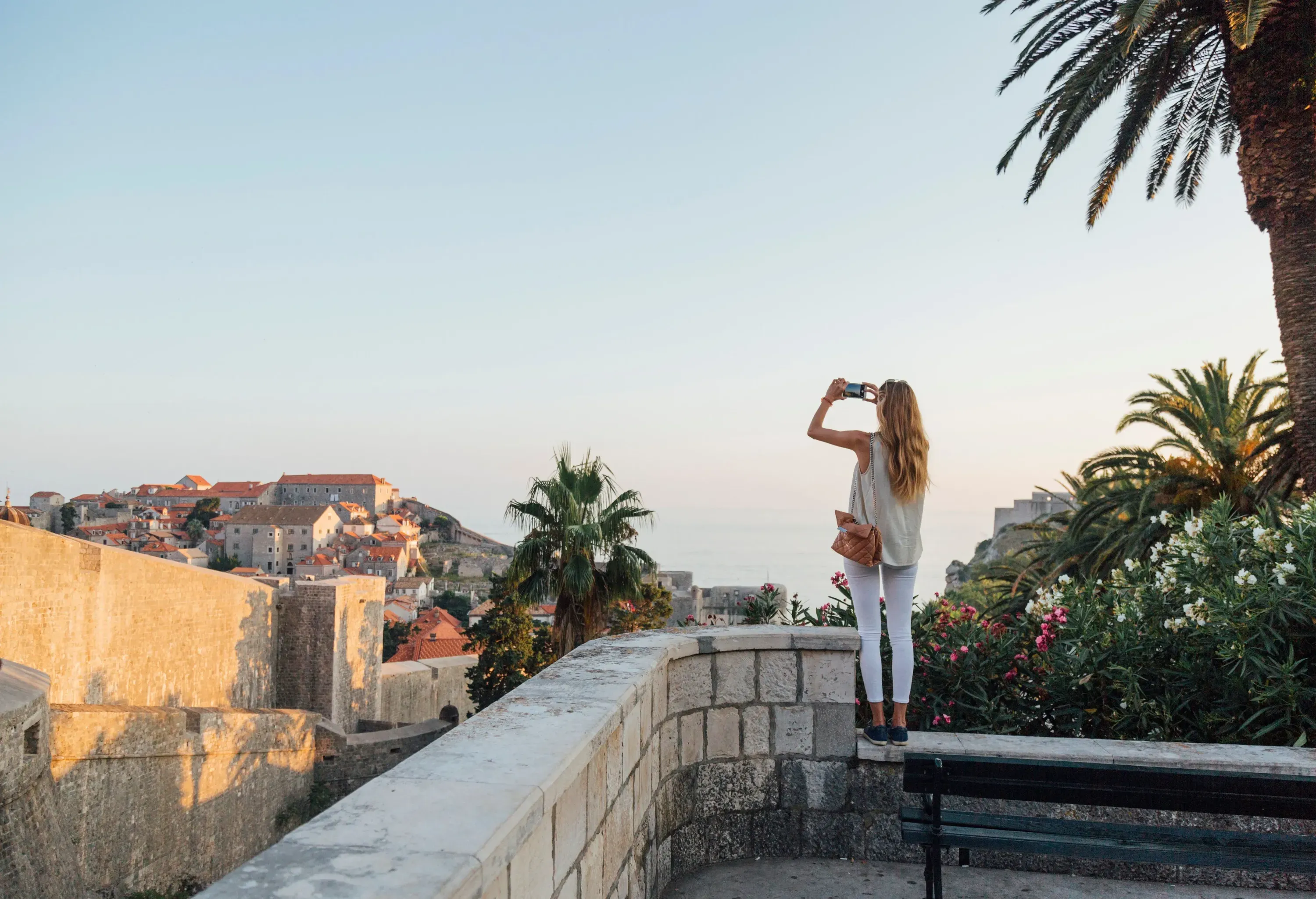 A long-haired blonde young lady takes a picture of a cityscape on a hill on her phone.