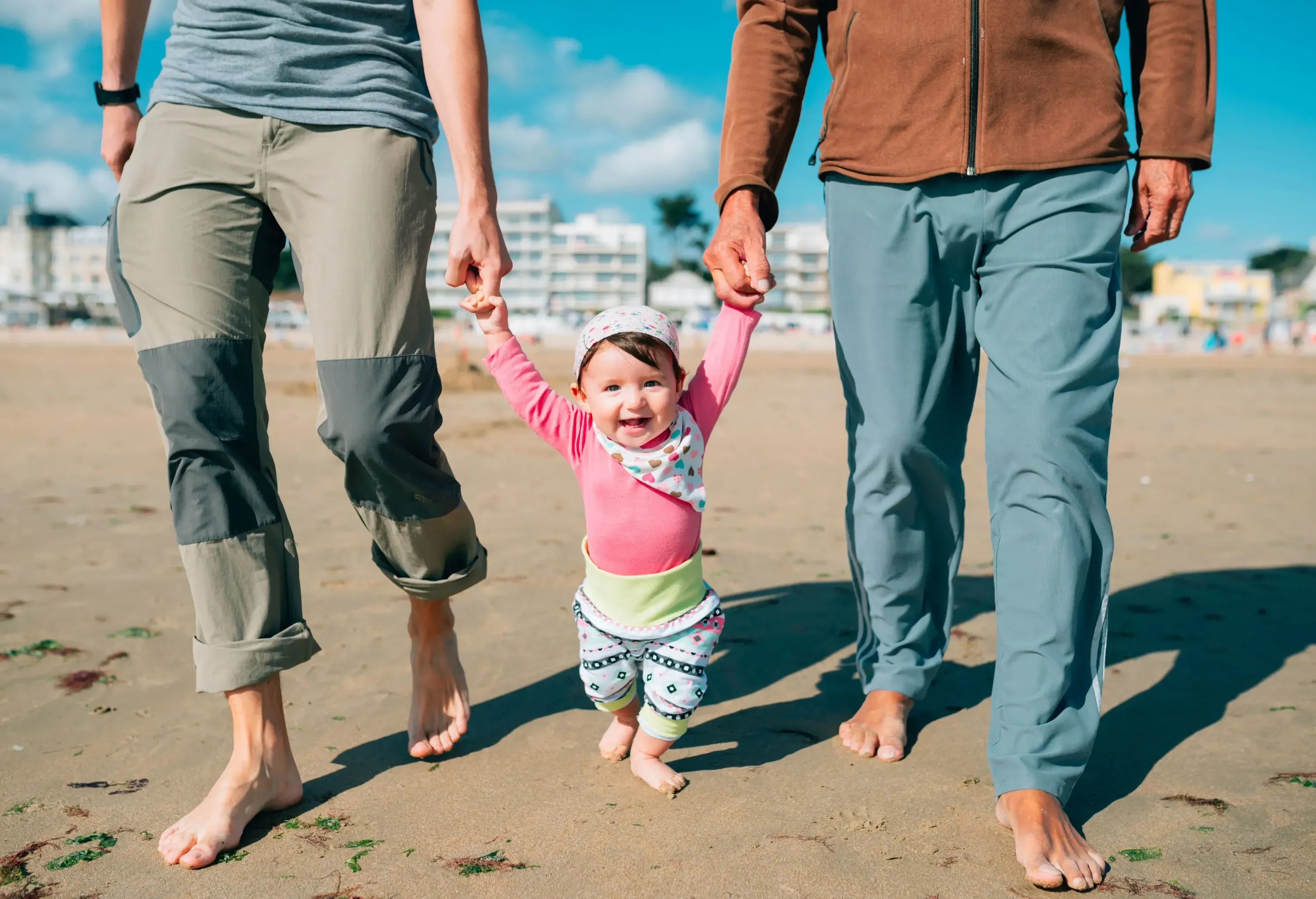 A baby girl wearing a turban and a bib is smiling while walking and being handheld by two adults on the sand.