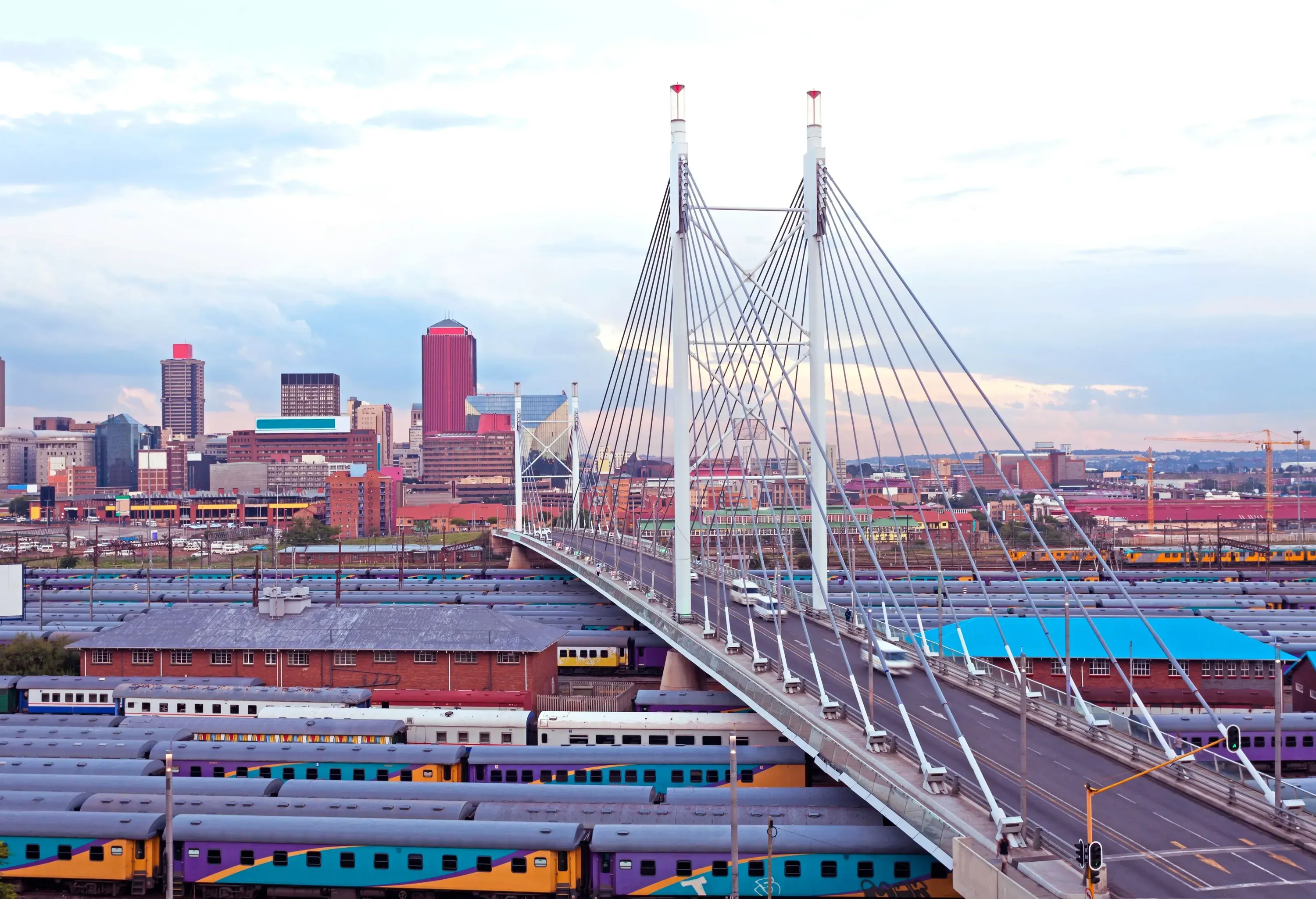 The iconic Nelson Mandela Bridge spans gracefully over a vibrant train park area, with an expansive urban skyline serving as a captivating backdrop.