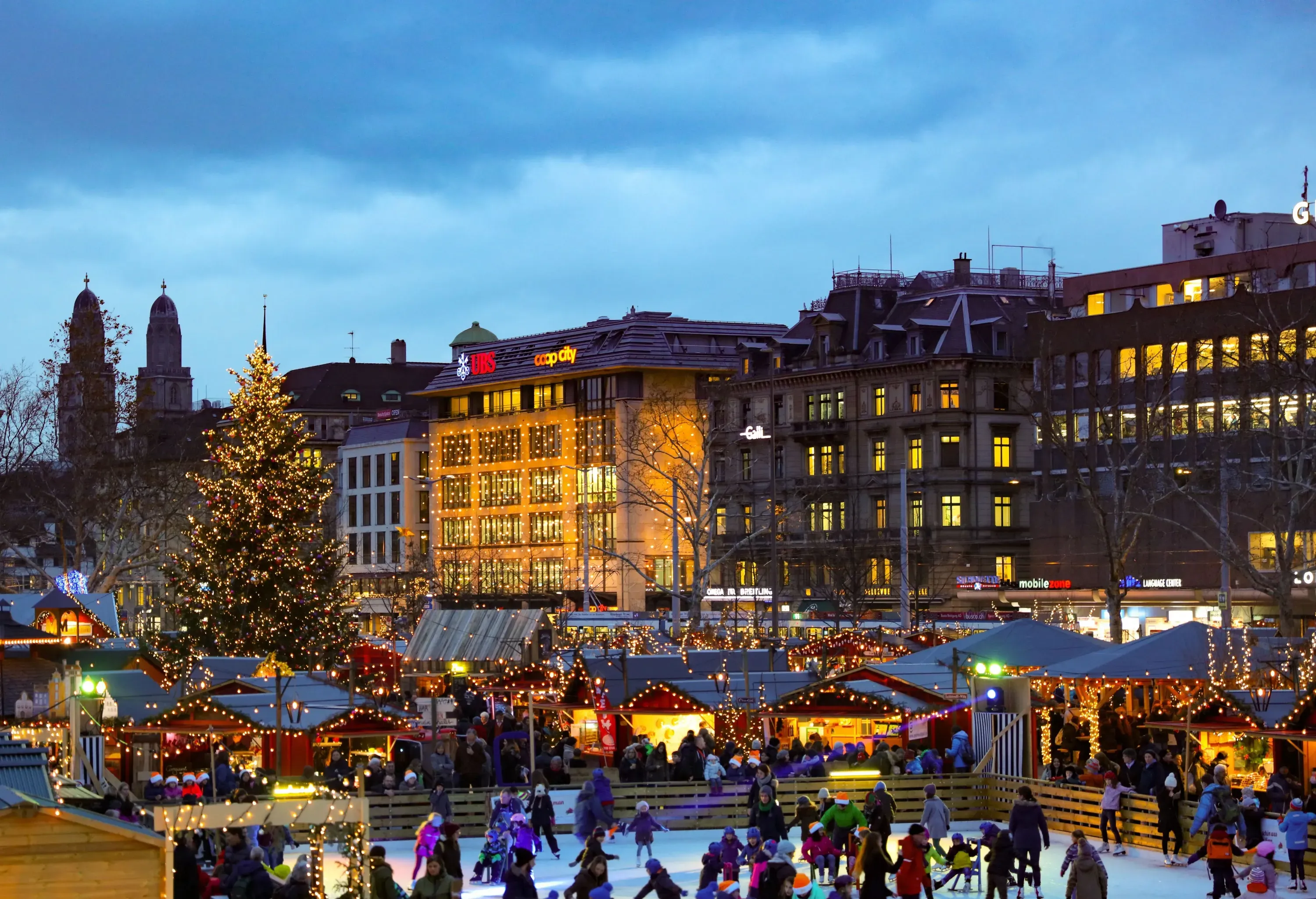 People are on a brightly lit Christmas market ice skating rink surrounded by classic buildings in the background.